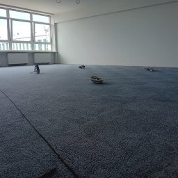 Jan Michurski Wykładziny Obiektowe - Niebieska wykładzina dywanowa z delikatnym wzorem w przestronnym, jasnym pomieszczeniu biurowym z dużymi oknami, widoczne zwinięte kable elektryczne i listwa przypodłogowa wzdłuż ściany.