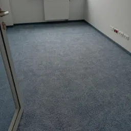 Pokój z nową, niebieską wykładziną dywanową o delikatnym wzorze, widoczne gniazdka elektryczne przy ścianie i fragment ramy drzwiowej.