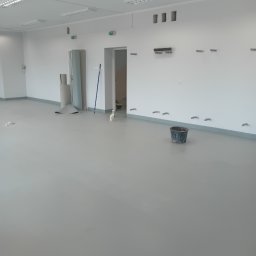 Jan Michurski Wykładziny Obiektowe - Szara wylewka samopoziomująca w surowym, oświetlonym wnętrzu biurowym, z widocznymi otworami w ścianach, zwiniętą rolką materiału pod ścianą i wiaderkiem na podłodze.