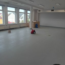 Jan Michurski Wykładziny Obiektowe - Duże, puste pomieszczenie z szarą, gładką podłogą, widoczne okna z grzejnikami, kolumna, wiadro i torba narzędziowa; przygotowane do dalszych prac wykończeniowych.