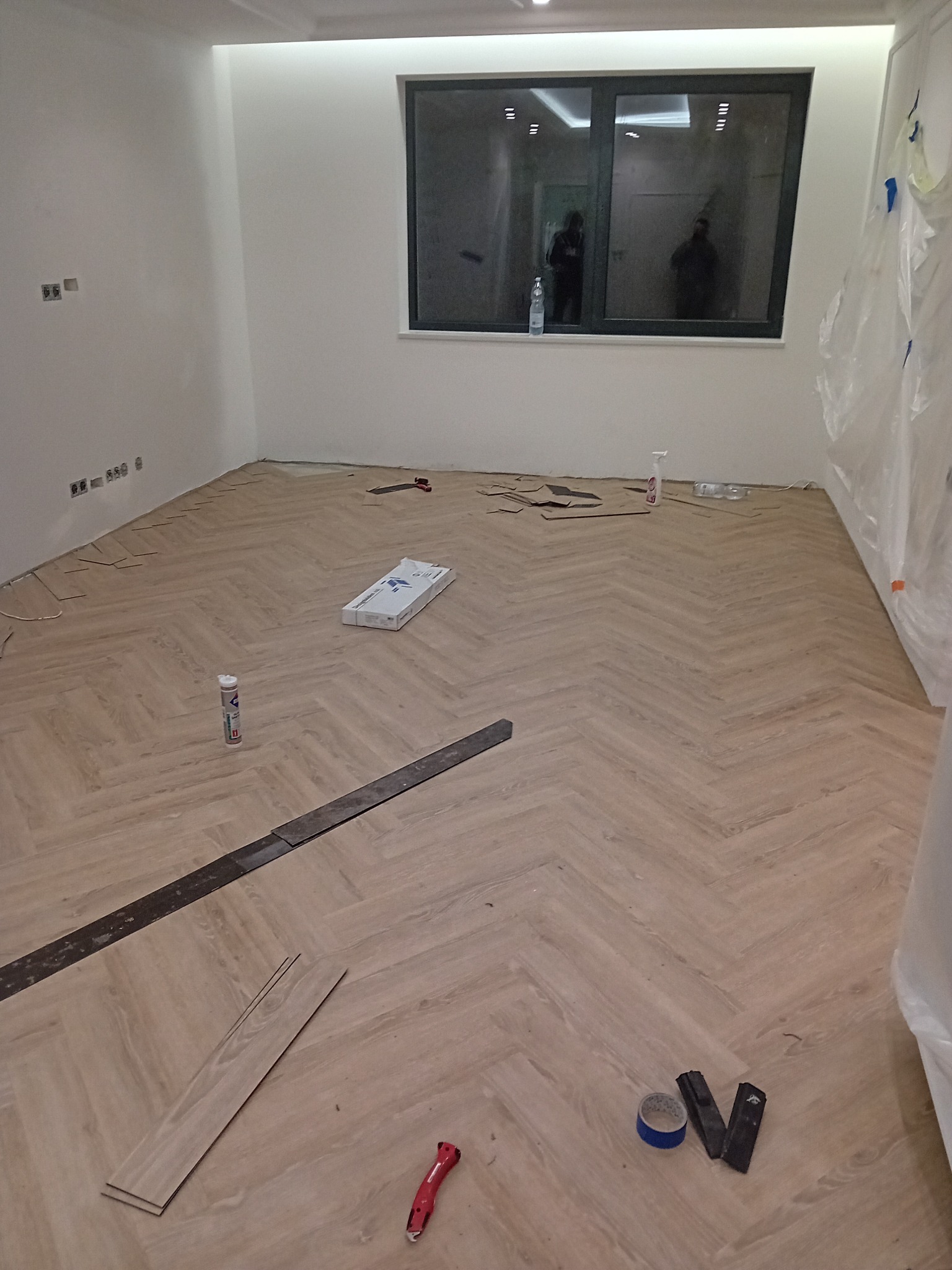 Układanie jasnych paneli w jodełkę francuską, widoczne narzędzia, listwy przypodłogowe jeszcze nie zamontowane, okno z odbiciami osób.