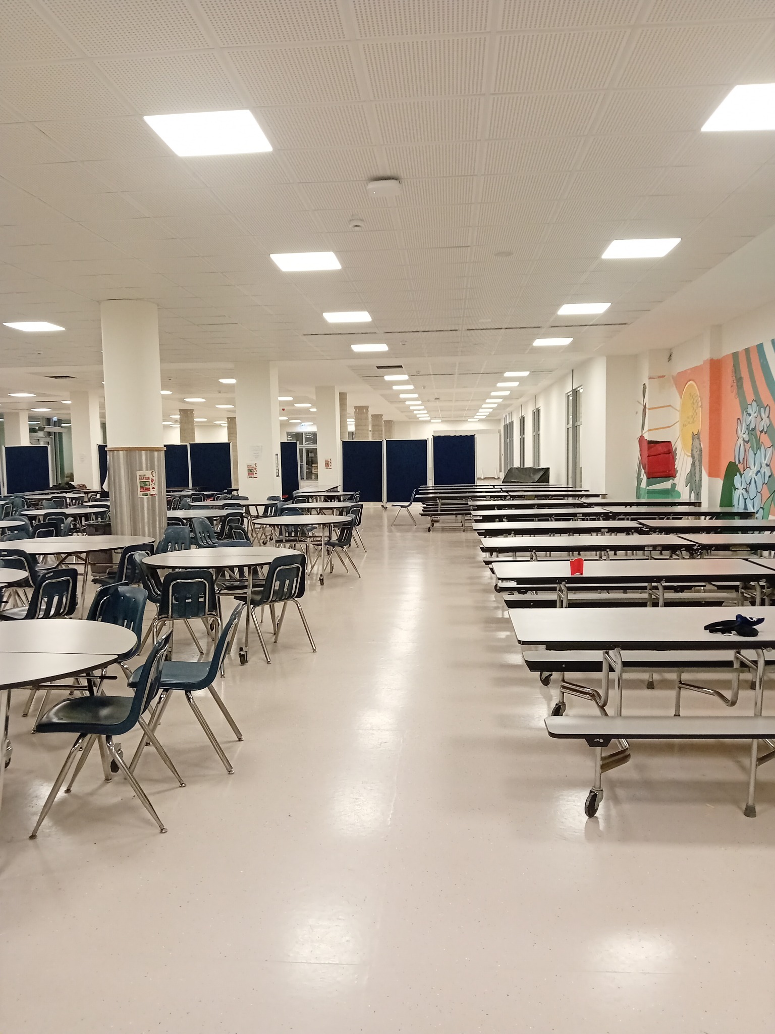 Rozległa stołówka z rzędami stołów i krzeseł, jasna podłoga z drobnymi plamkami, białe kolumny i sufit z panelami, w tle widoczny mural na ścianie.