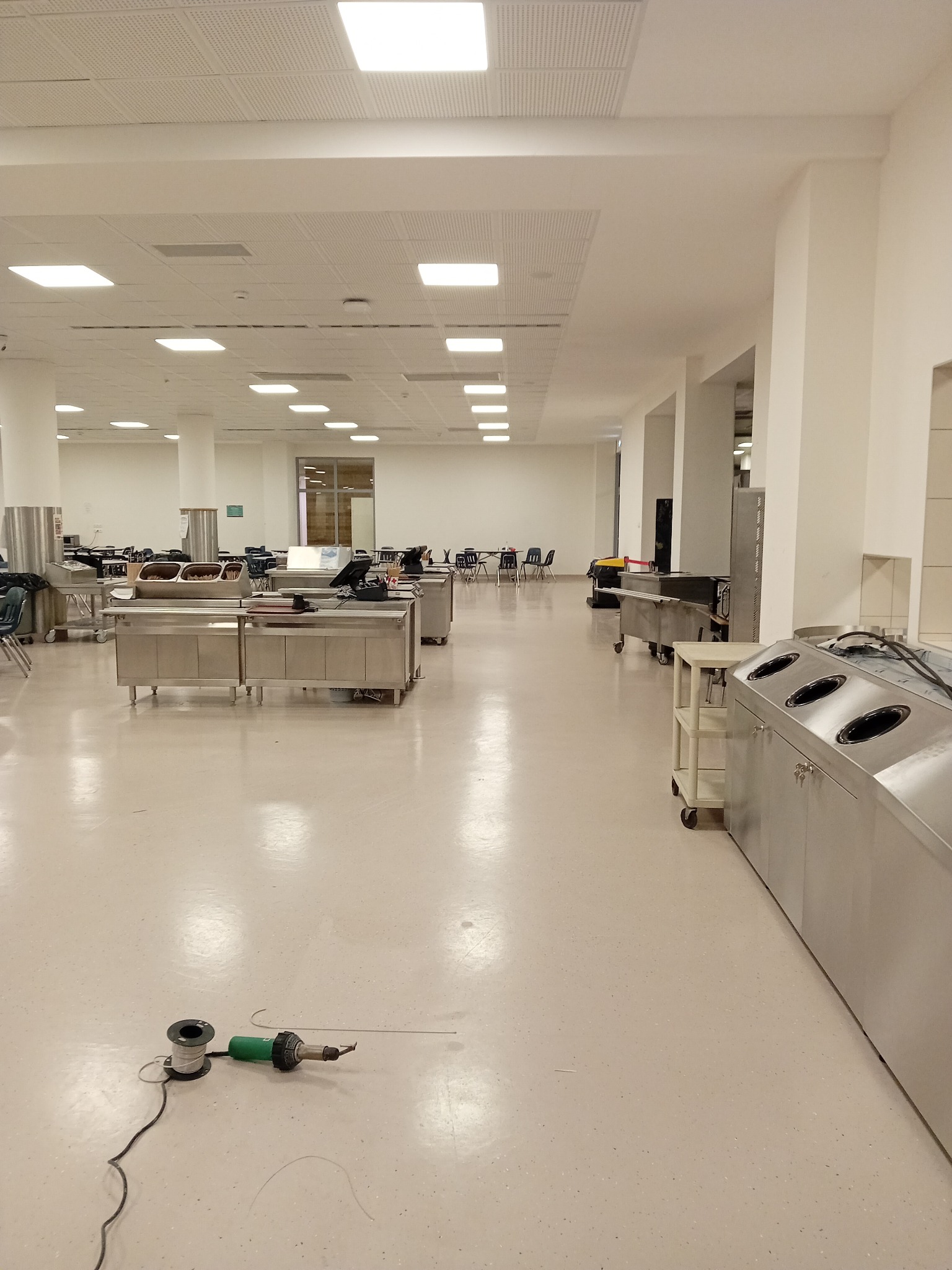 Duża, pusta stołówka z nową, jasną podłogą z tworzywa sztucznego, na której leży porzucone narzędzie do spawania wykładzin.