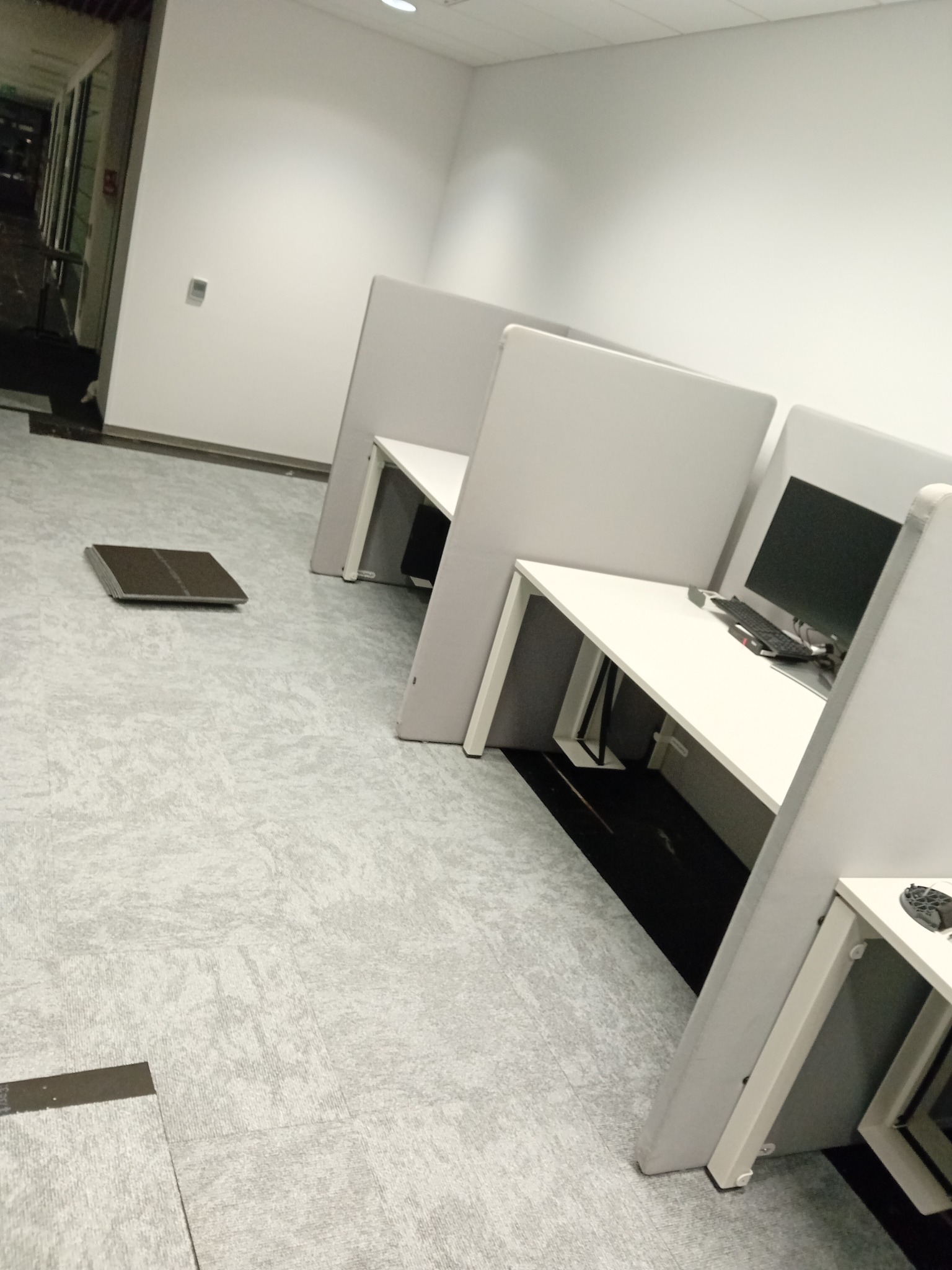 Układanie jasnoszarej wykładziny dywanowej w biurze z białymi biurkami i ściankami działowymi, widoczna niedokończona krawędź i stos płytek wykładzinowych.