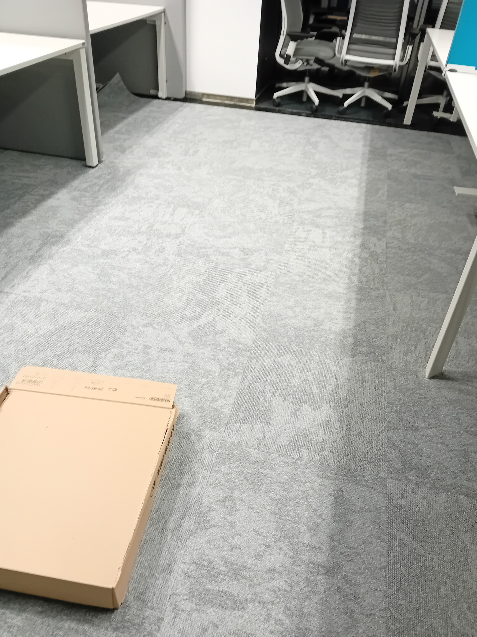 Szara wykładzina dywanowa w biurze, z kartonem na podłodze i fragmentami biurek oraz krzeseł w tle.