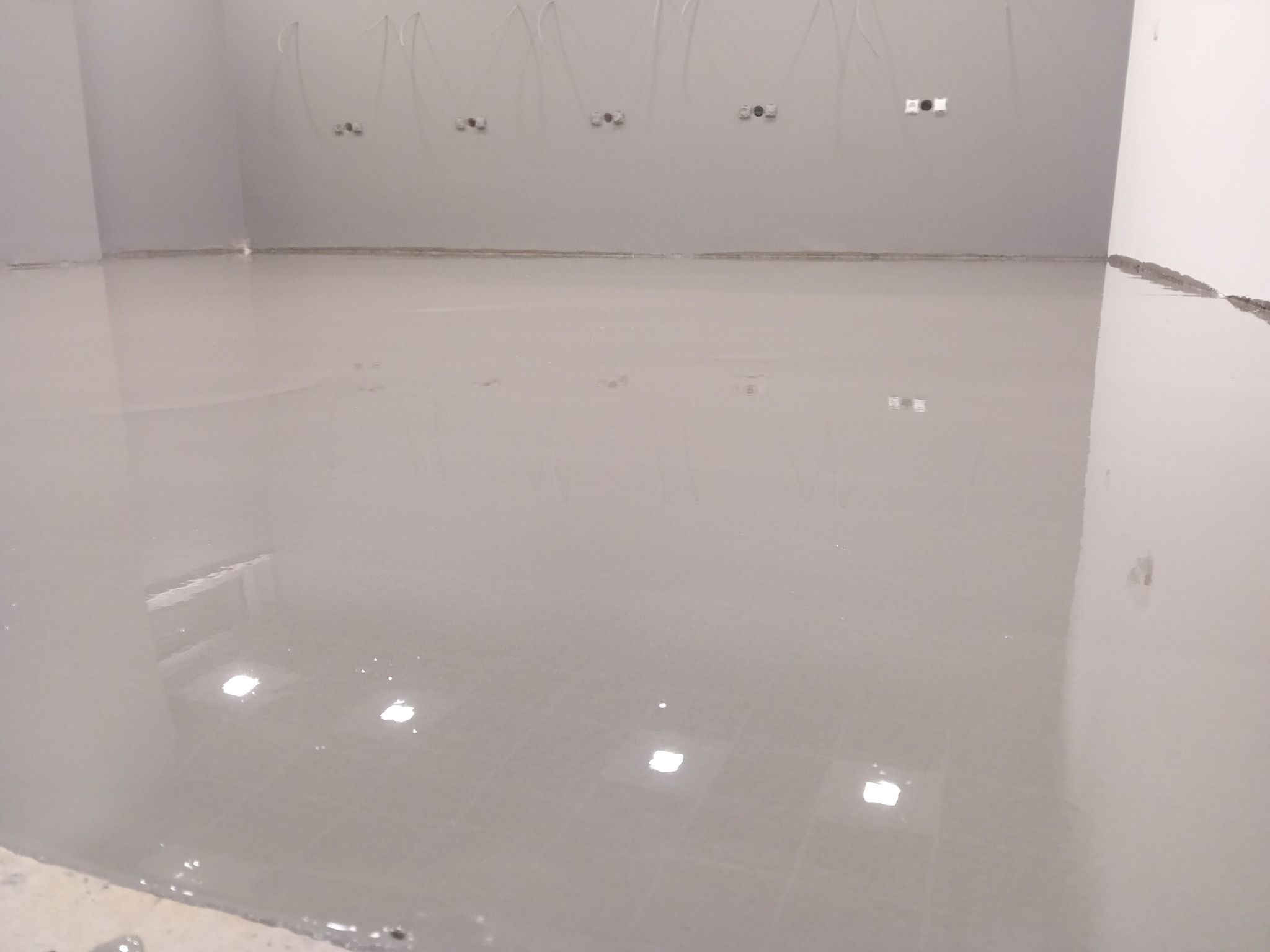 Wylany, samopoziomujący podkład podłogowy w szarym odcieniu, widoczne przewody elektryczne wystające ze ściany.