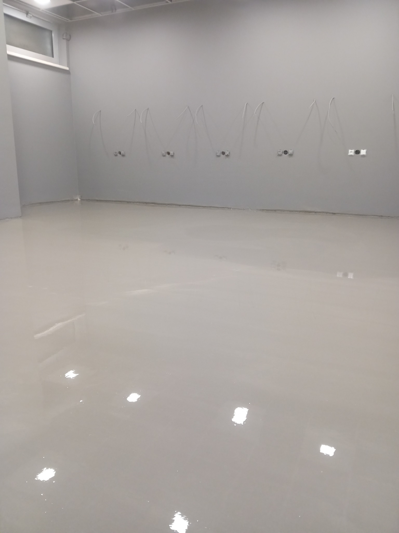 Świeżo wylana, samopoziomująca, szara posadzka żywiczna w pustym pomieszczeniu z wystającymi kablami ze ściany.