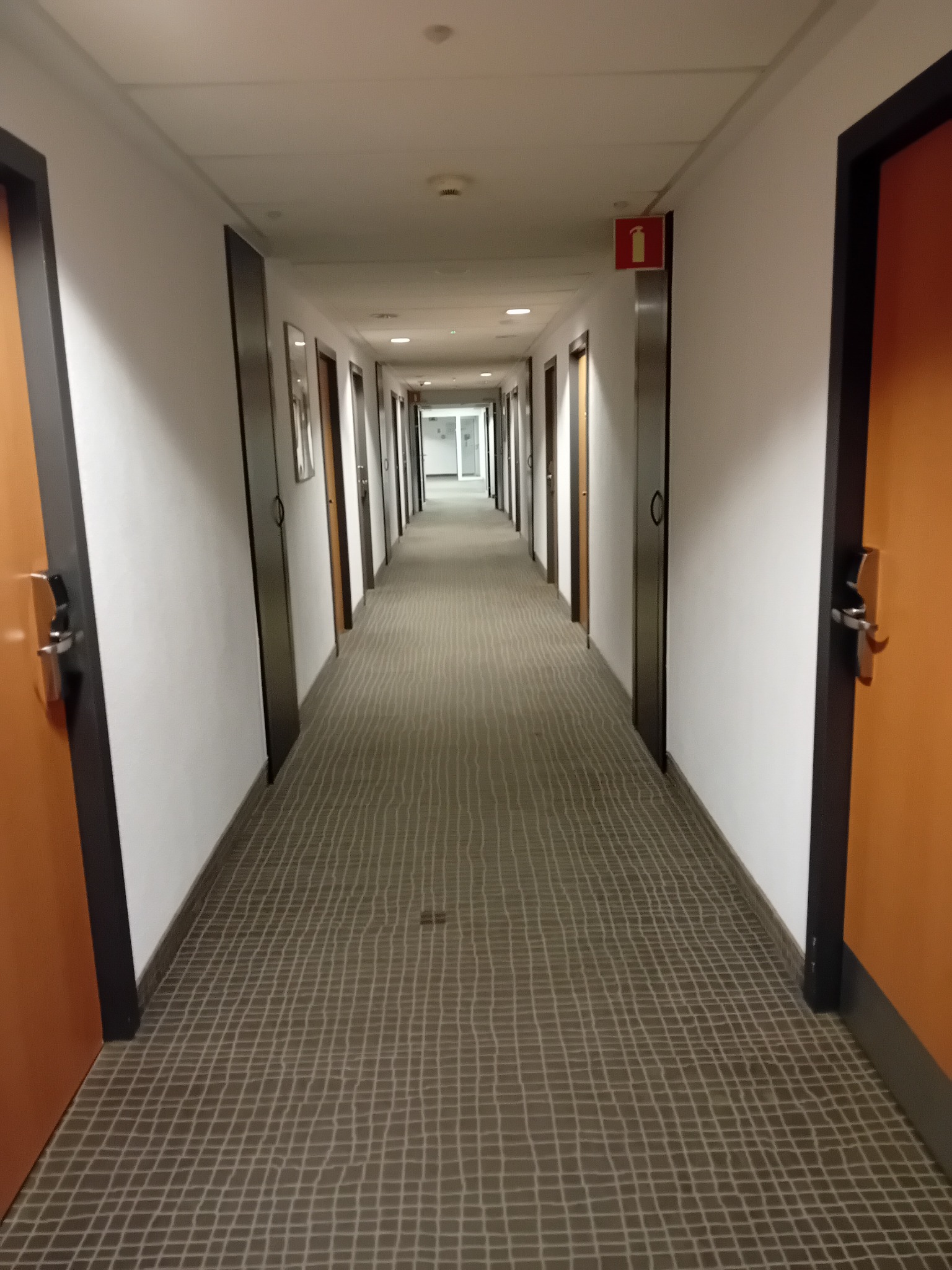 Długi korytarz hotelowy z szarą wykładziną w kratę, pomarańczowe drzwi po obu stronach, widoczny znak przeciwpożarowy na końcu korytarza.