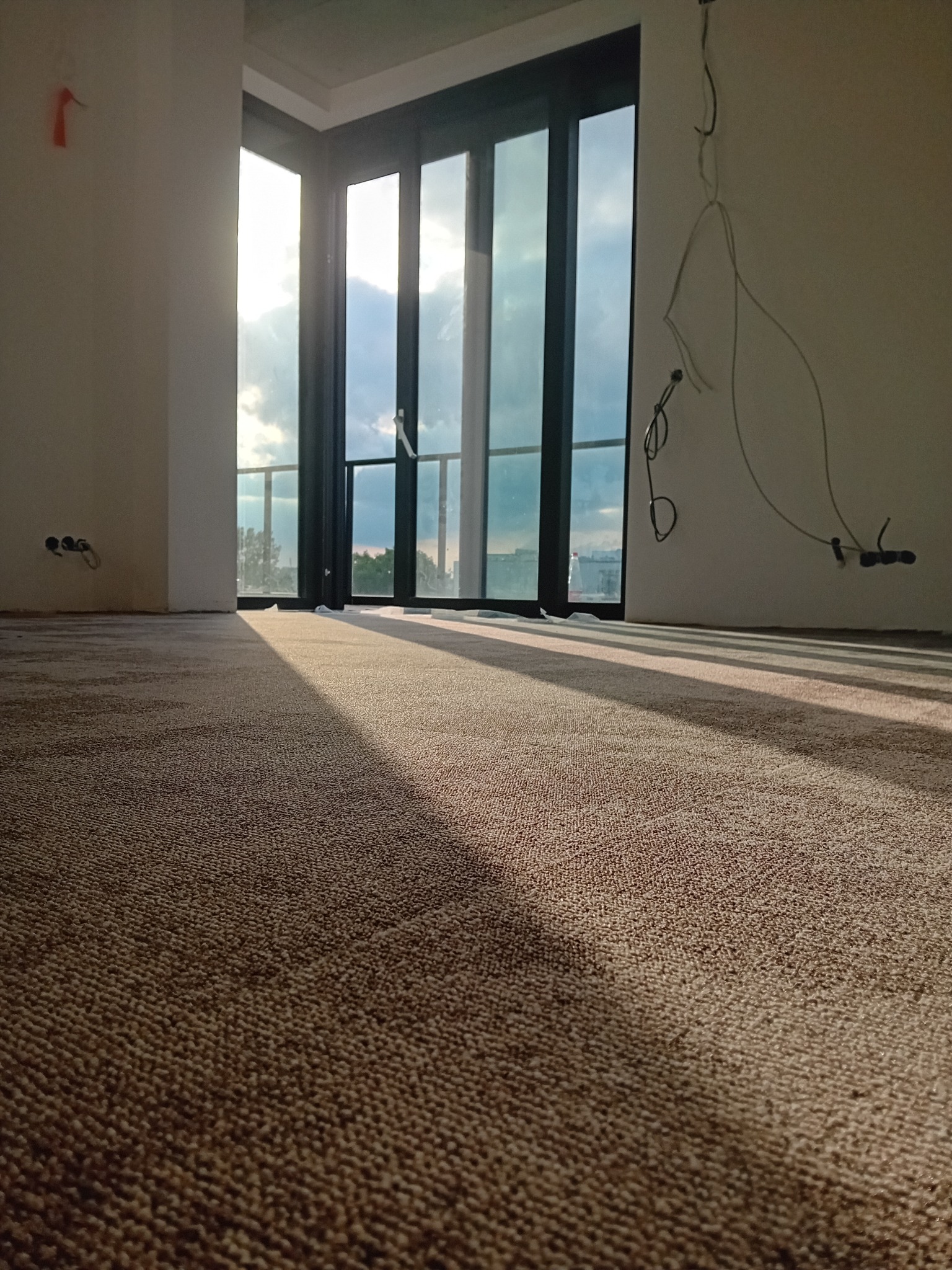 Beżowa wykładzina dywanowa w nowym pomieszczeniu z dużym oknem balkonowym, wpadające promienie słoneczne tworzą geometryczne cienie na podłodze, widoczne niedokończone instalacje elektryczne...