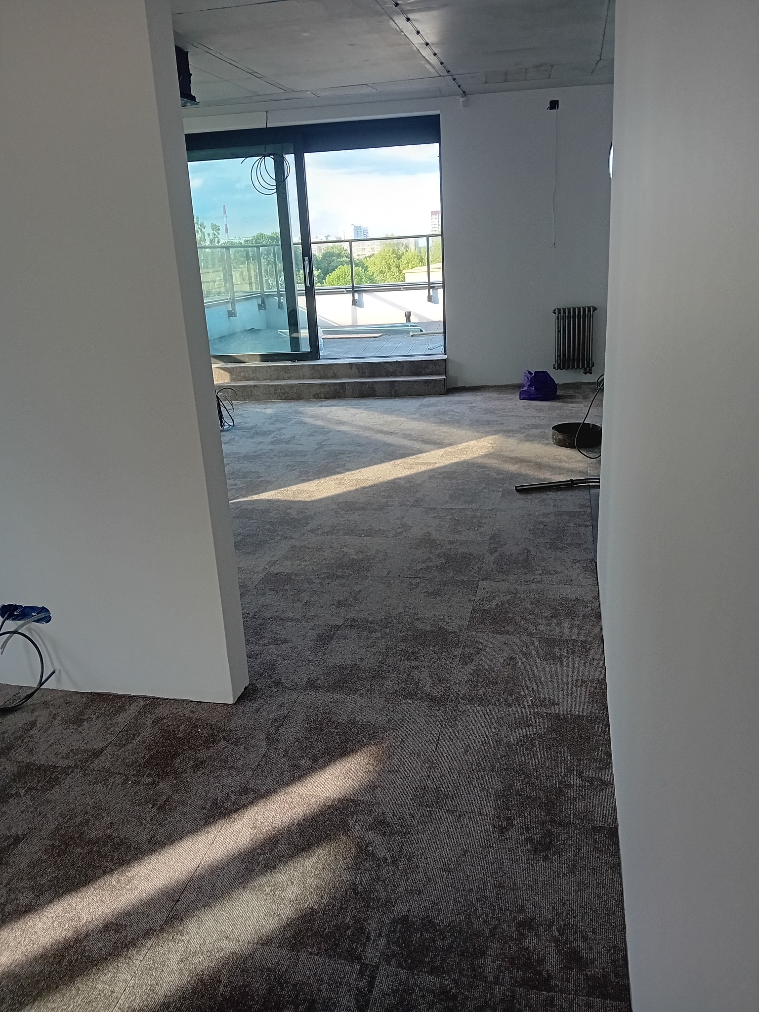 Nowo ułożona, szara wykładzina dywanowa w jasnym pomieszczeniu z wyjściem na balkon, widoczne narzędzia i niedokończone prace wykończeniowe.