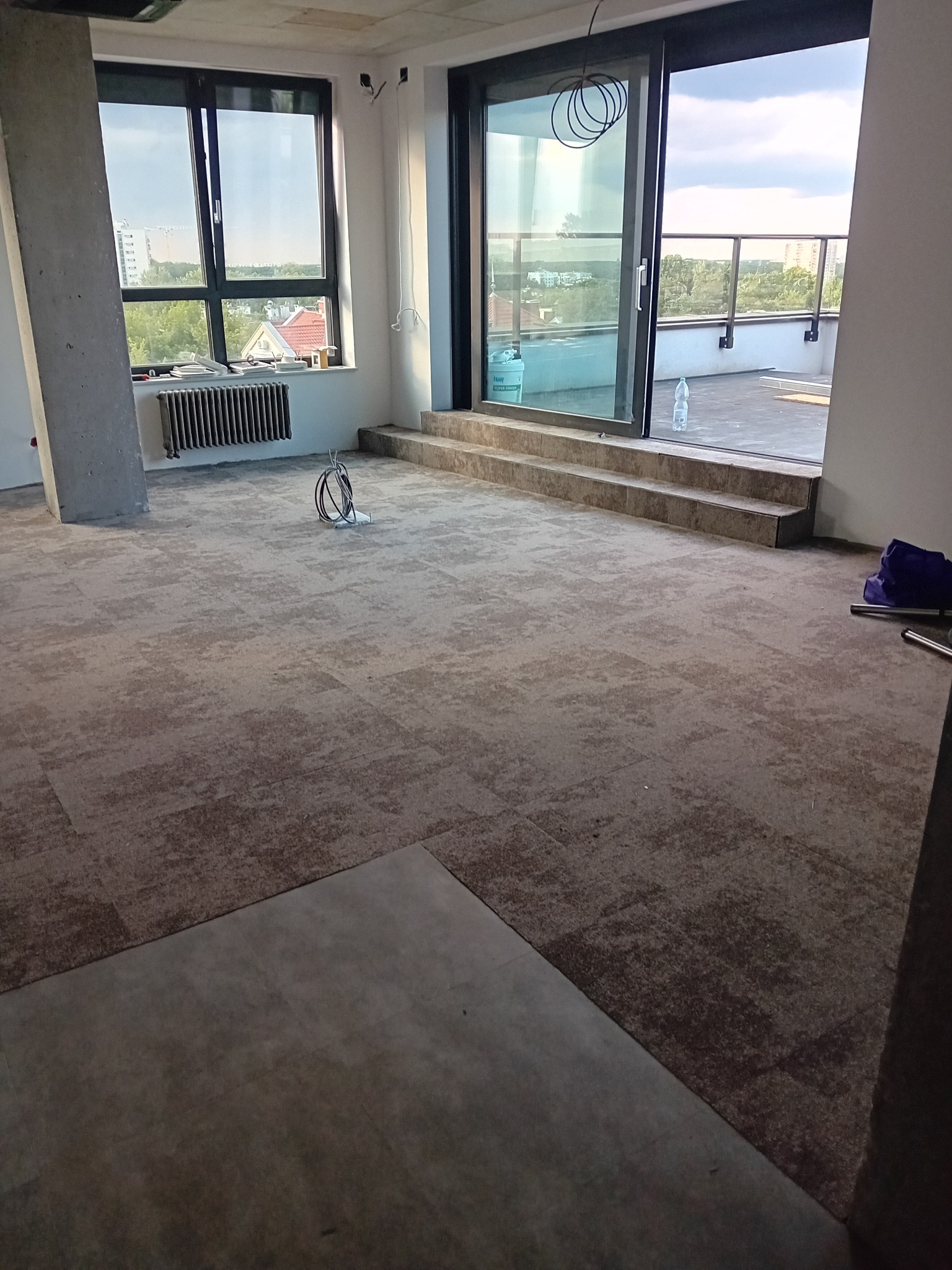 Wnętrze w trakcie wykańczania z ułożoną wykładziną dywanową w neutralnych odcieniach, widoczne fragmenty instalacji elektrycznej, drzwi balkonowe z widokiem na miasto i niezabezpieczone krawędzie...