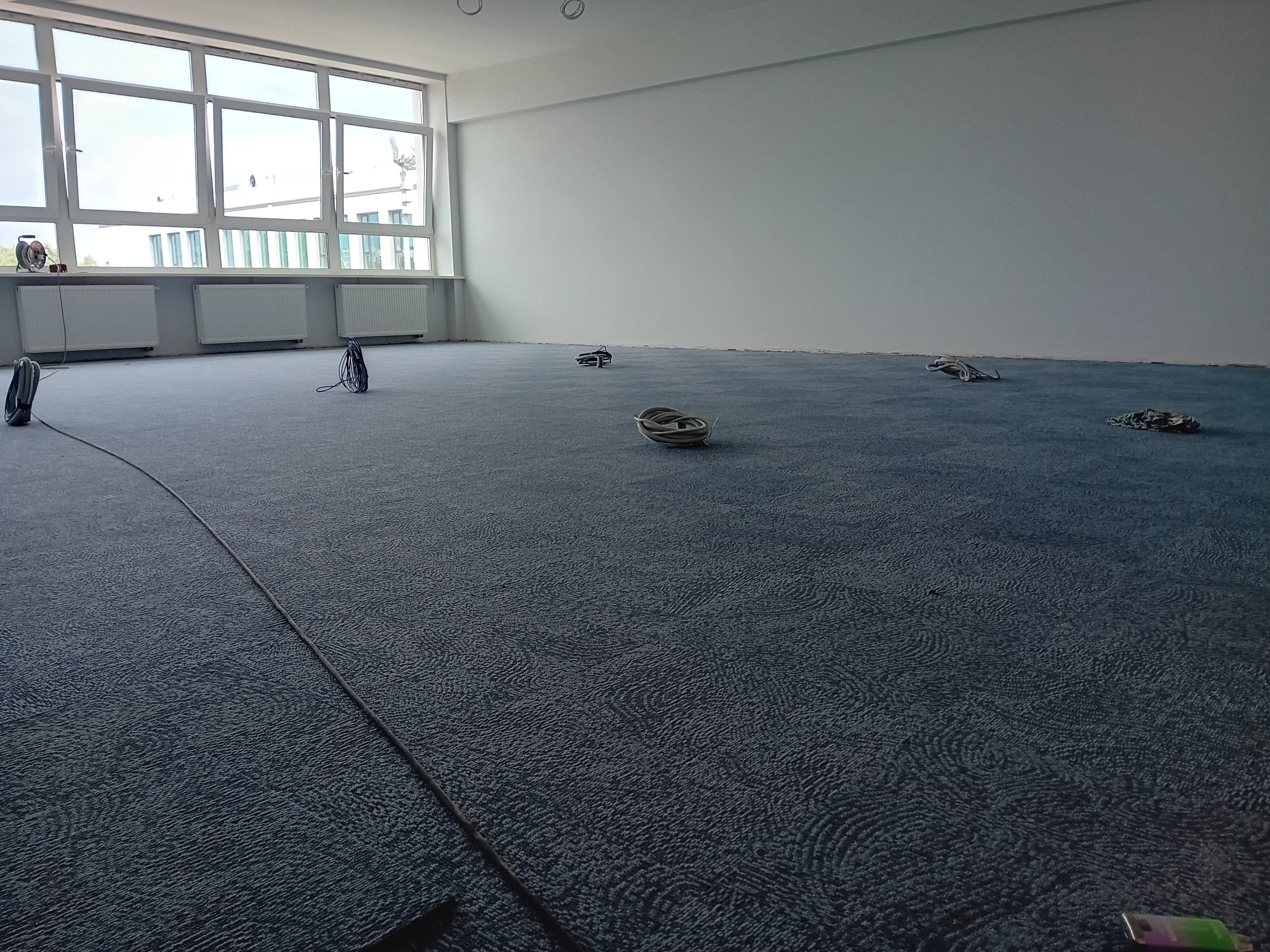 Niebieska wykładzina dywanowa z delikatnym wzorem w przestronnym, jasnym pomieszczeniu biurowym z dużymi oknami, widoczne zwinięte kable elektryczne i listwa przypodłogowa wzdłuż ściany.