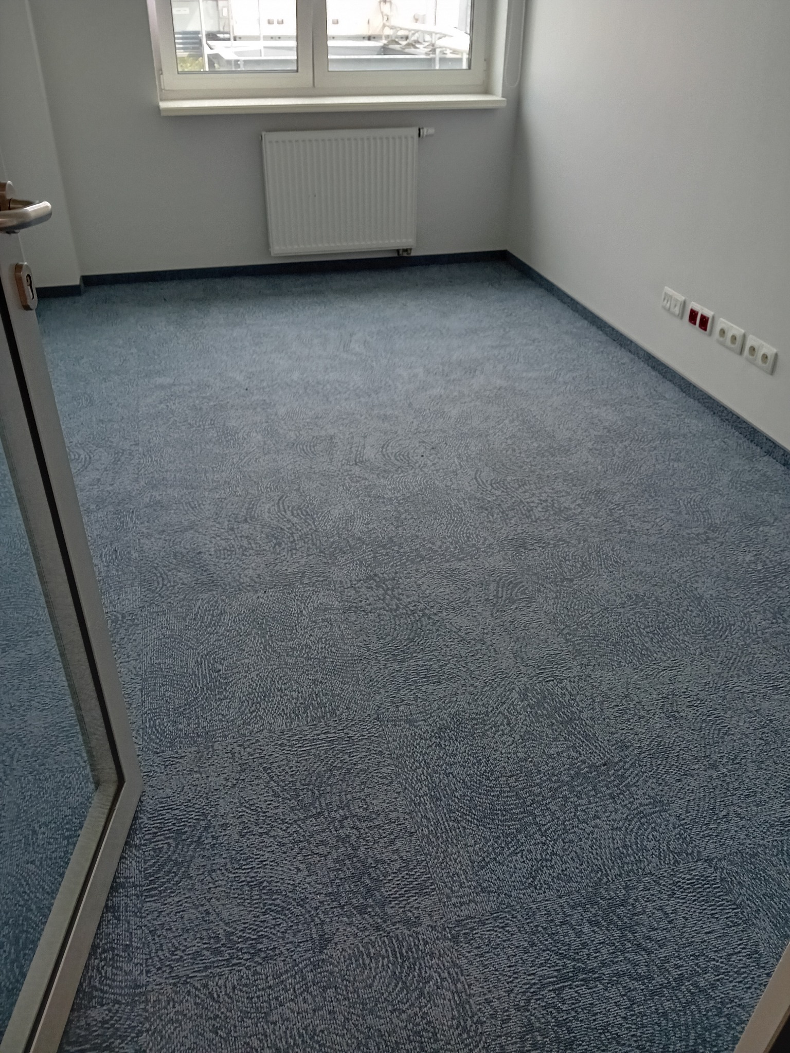 Pokój z nową, niebieską wykładziną dywanową o delikatnym wzorze, widoczne gniazdka elektryczne przy ścianie i fragment ramy drzwiowej.