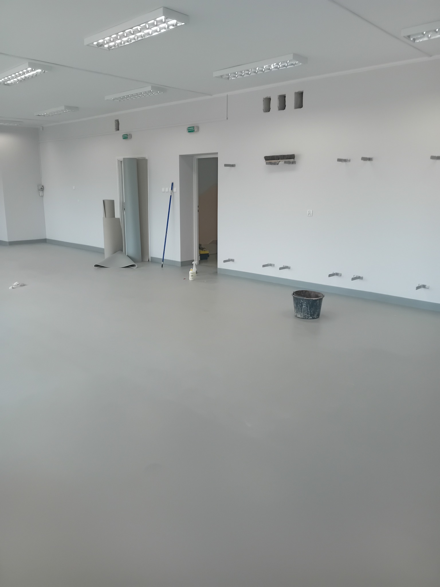 Szara wylewka samopoziomująca w surowym, oświetlonym wnętrzu biurowym, z widocznymi otworami w ścianach, zwiniętą rolką materiału pod ścianą i wiaderkiem na podłodze.