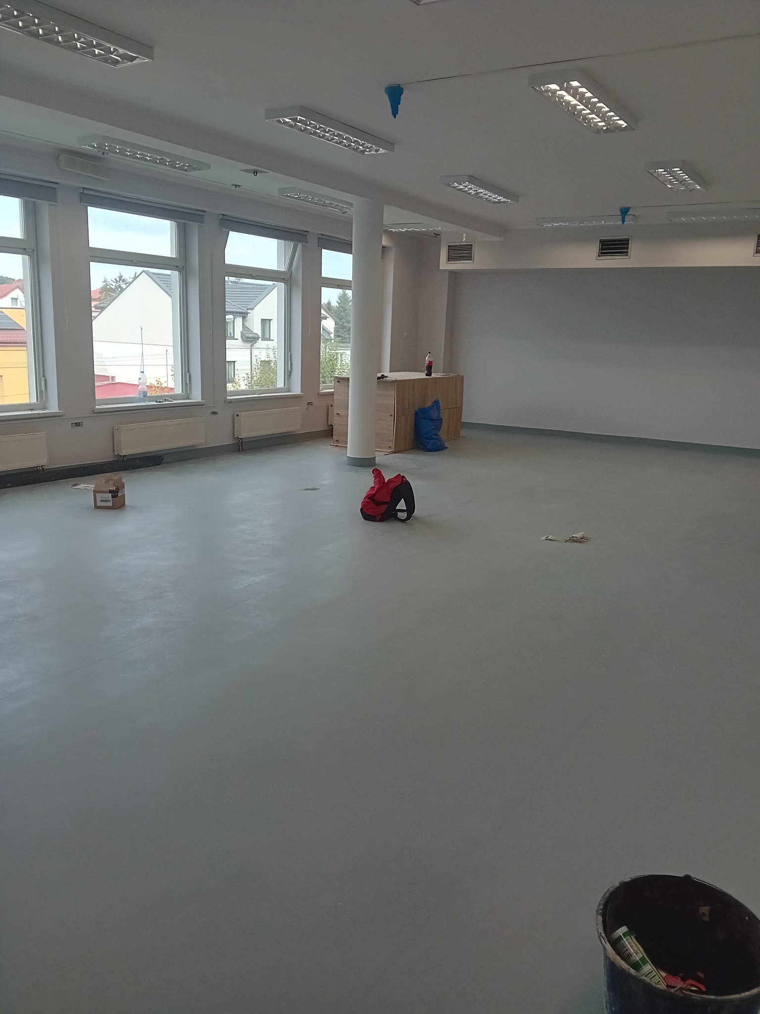 Duże, puste pomieszczenie z szarą, gładką podłogą, widoczne okna z grzejnikami, kolumna, wiadro i torba narzędziowa; przygotowane do dalszych prac wykończeniowych.