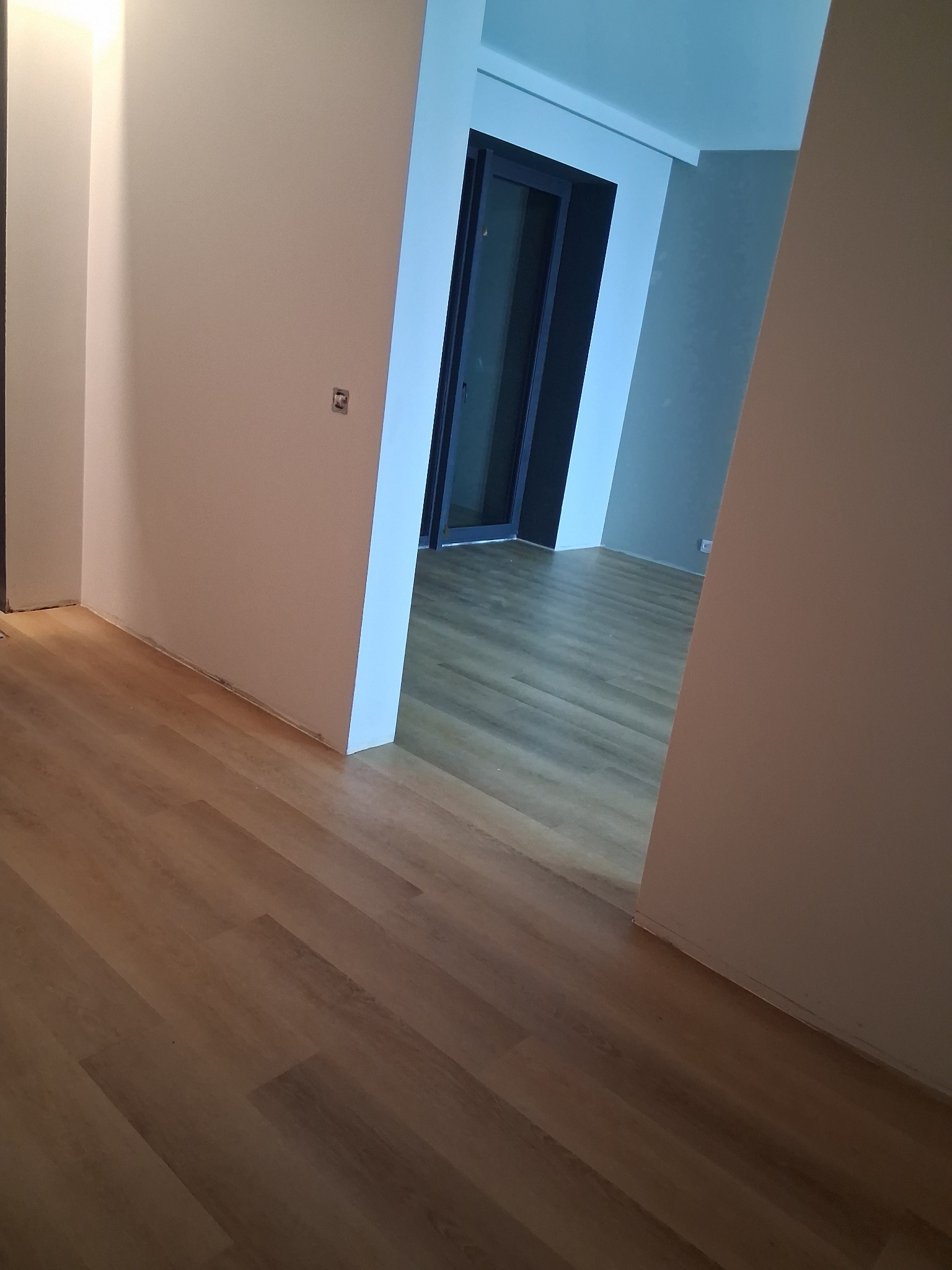 Ukończone wnętrze z panelami podłogowymi w odcieniach brązu, widoczne łączenia między panelami, ściany pomalowane na biało i szaro, widoczne drzwi w tle.