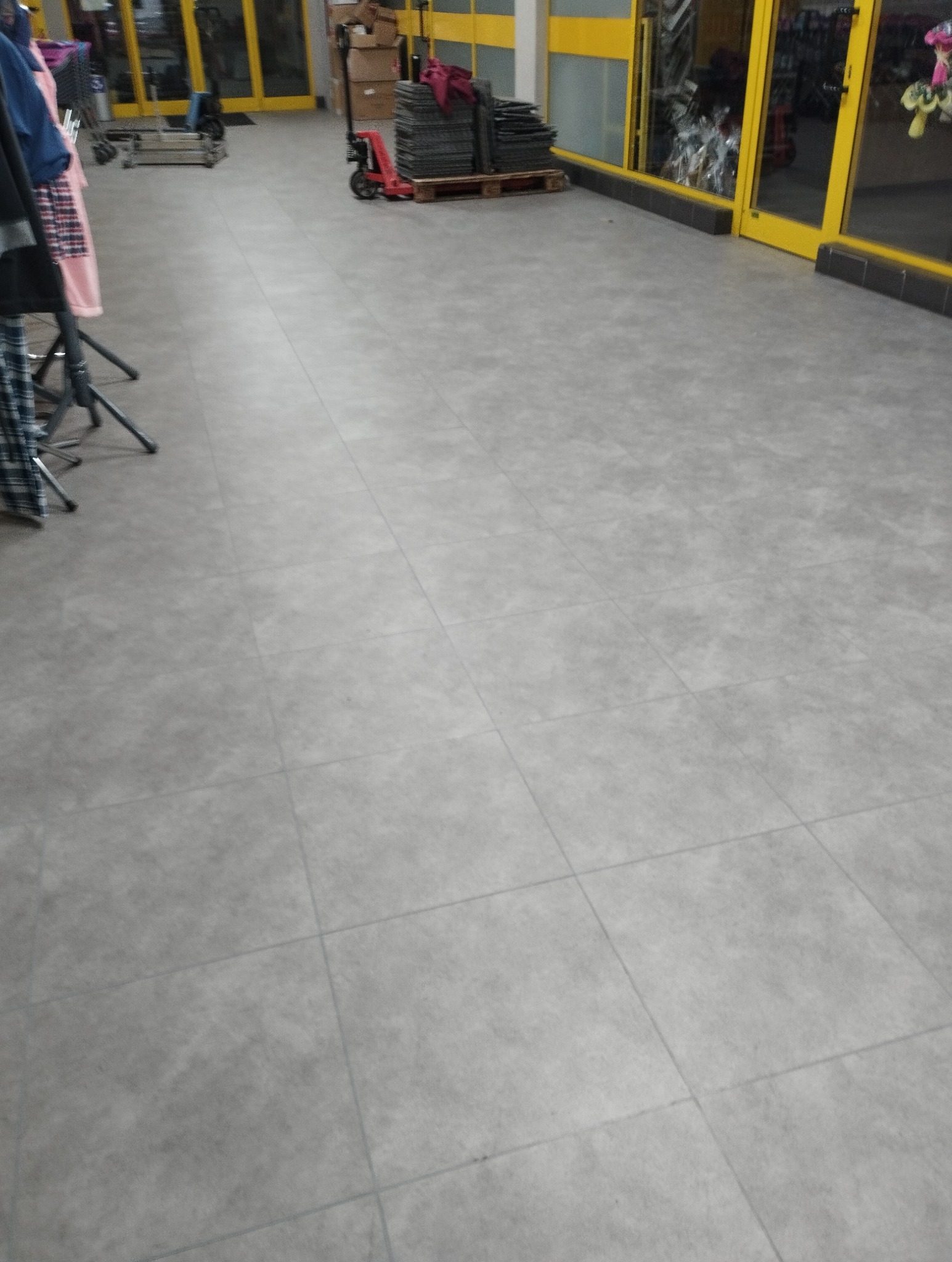 Szare płytki podłogowe ułożone w regularny wzór, z widocznymi fugami, na tle sklepowego wnętrza z żółtymi ramami drzwi i paletą z płytkami na wózku paletowym.