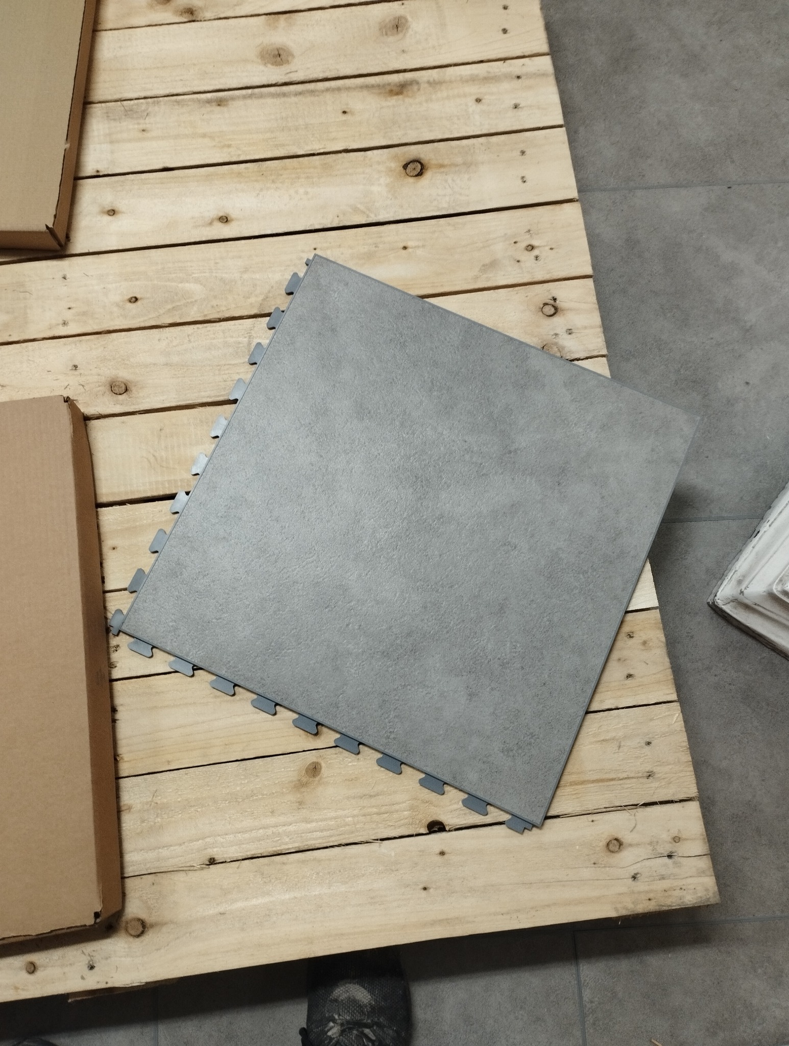 Szara płyta podłogowa z wypustkami do łączenia, leżąca na drewnianej palecie, obok kartonowe pudełko.