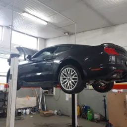 Czarny samochód sportowy, prawdopodobnie Ford Mustang, podniesiony na podnośniku w warsztacie samochodowym, z otwartą maską, widoczne narzędzia i wyposażenie warsztatu.