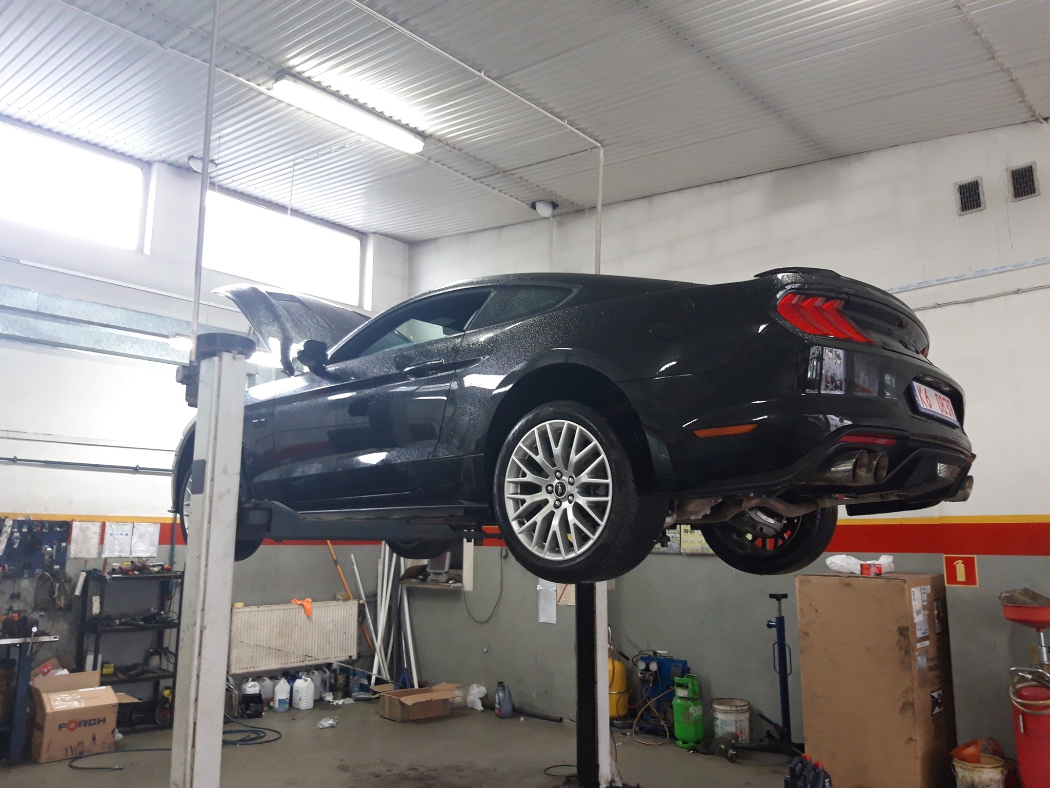Czarny samochód sportowy, prawdopodobnie Ford Mustang, podniesiony na podnośniku w warsztacie samochodowym, z otwartą maską, widoczne narzędzia i wyposażenie warsztatu.
