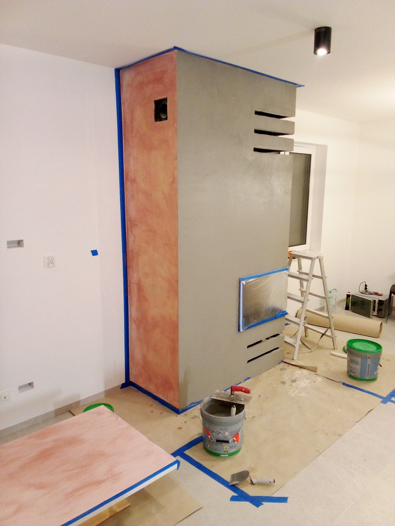 Wnętrze w trakcie prac wykończeniowych: obudowa kominka z szarą gładzią i dekoracyjną ścianką pokrytą tynkiem imitującym marmur, zabezpieczona taśmą malarską, drabina i narzędzia na podłodze.