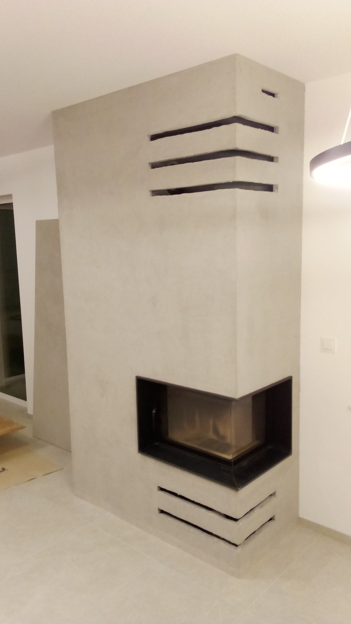 Nowoczesny, narożny kominek z szarego betonu architektonicznego, z poziomymi szczelinami wentylacyjnymi, w jasnym wnętrzu, po remoncie.