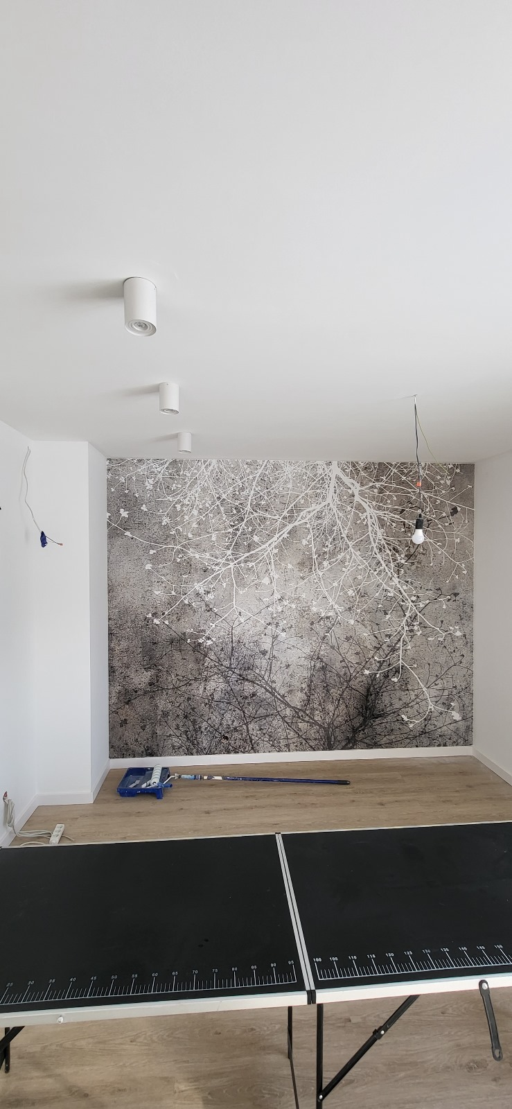 Pokój po malowaniu z fototapetą z motywem drzew, widoczny wałek do malowania i stół do tenisa stołowego.