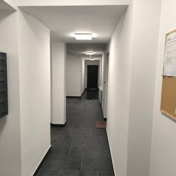 ELMAL - Korytarz w budynku wielorodzinnym z czarną podłogą z płytek i białymi ścianami. Widoczne skrzynki pocztowe i tablica ogłoszeń.