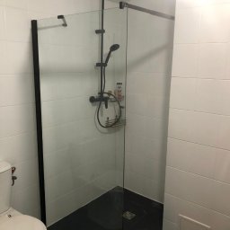 ELMAL - Nowoczesna kabina prysznicowa z czarną armaturą i szklaną ścianką w wyremontowanej łazience z białymi płytkami i ciemną podłogą. Widoczny fragment toalety.