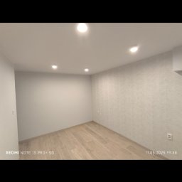 ELMAL - Jasne wnętrze pokoju po remoncie: parkiet, ściana z delikatnym wzorem, oświetlenie sufitowe. Widoczny fragment gniazdka elektrycznego. Ujęcie z narożnika.