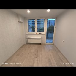 ELMAL - Jasne panele podłogowe w pustym pokoju z oknem balkonowym i grzejnikiem pod oknem. Ściana z geometrycznym wzorem. Widok na zewnątrz z zielenią.