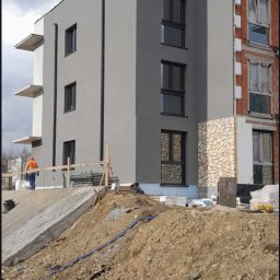 ELMAL - Wielopiętrowy budynek po częściowym malowaniu elewacji na szaro, z fragmentami w stanie surowym i osobą w oddali. Widoczne prace ziemne.
