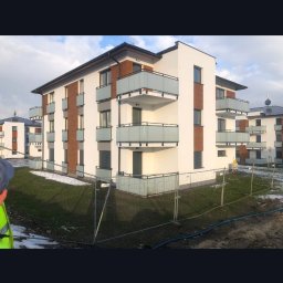 ELMAL - Nowoczesny, trzypiętrowy budynek mieszkalny z balkonami i elementami drewnopodobnymi na elewacji, otoczony trawnikiem i tymczasowym ogrodzeniem.