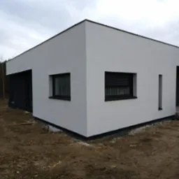 Nowoczesny, parterowy dom z płaskim dachem, wykończony białą elewacją i czarnymi detalami, takimi jak obramowania okien i drzwi, widoczny fragment terenu przed budynkiem z niezagospodarowaną ziemią.