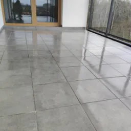 Błyszczące, szare płytki ceramiczne na podłodze balkonu z przeszkloną balustradą i widokiem na drzewa; w tle widoczne okno balkonowe z odbijającą się sylwetką osoby.