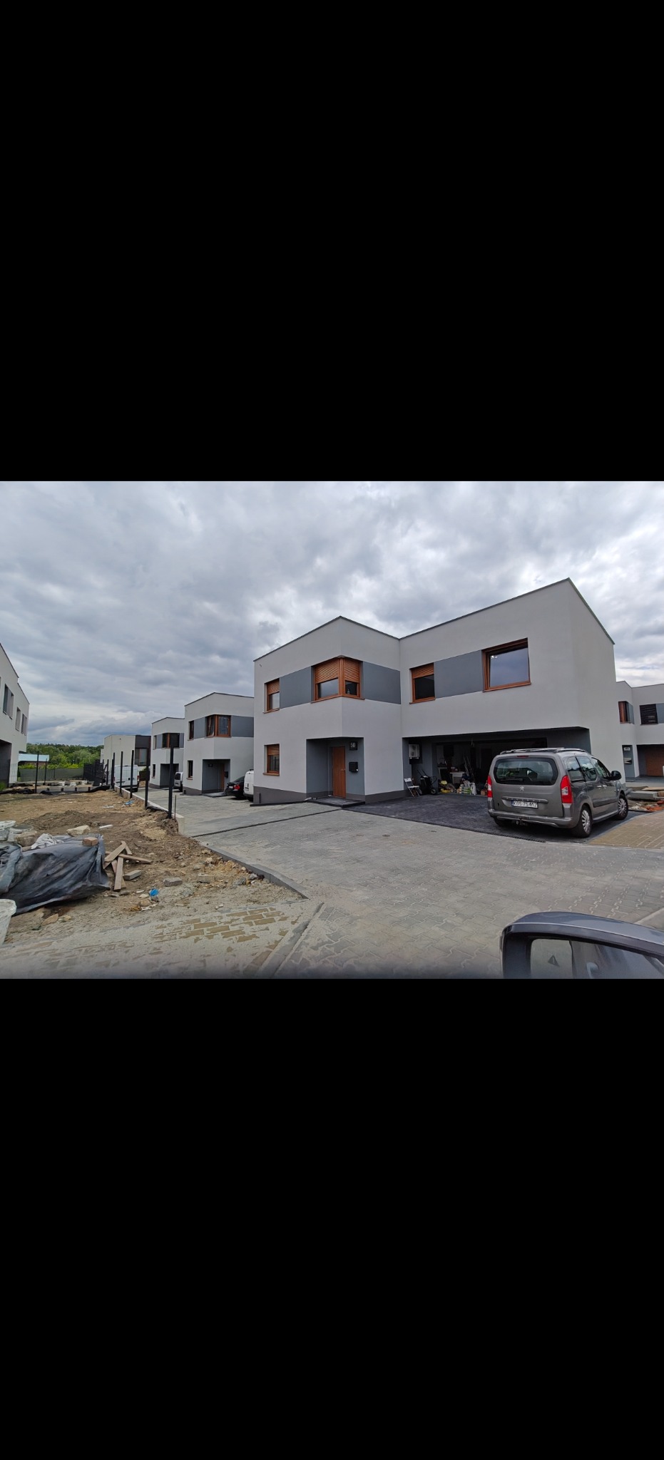 Nowoczesne, szeregowe domy z szarą elewacją i drewnianymi oknami w pochmurny dzień. Częściowo wykończony chodnik z kostki. Samochód na podjeździe.