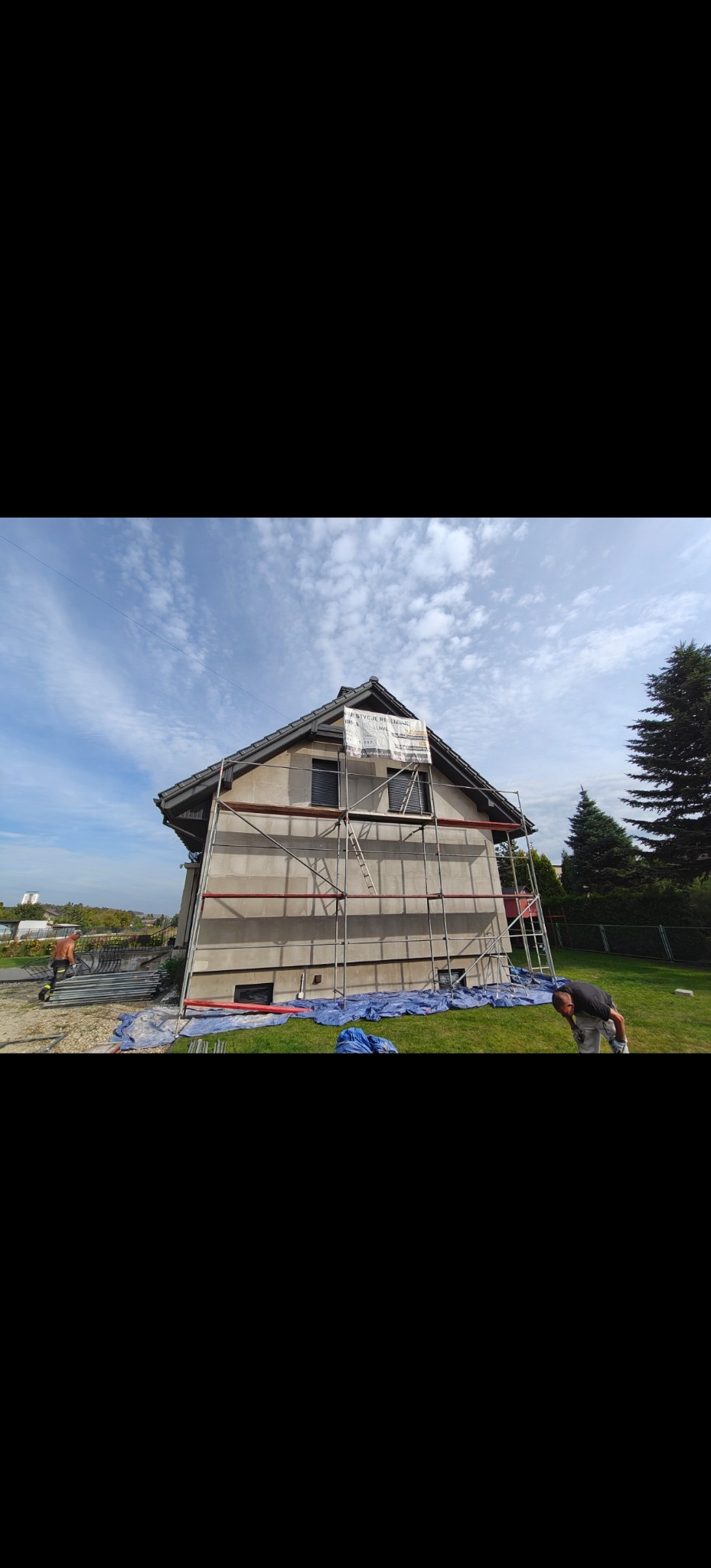 Dom jednorodzinny w trakcie prac elewacyjnych, otoczony rusztowaniem. Widoczni pracownicy i zabezpieczenia. Ujęcie w słoneczny dzień.