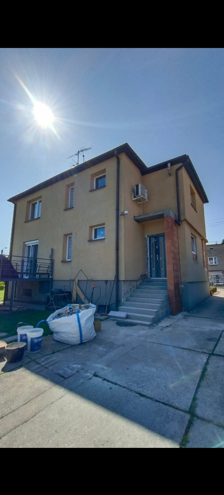 Dwupiętrowy dom z beżową elewacją w trakcie prac wykończeniowych. Widoczne worki z materiałami budowlanymi, schody wejściowe i fragment chodnika.