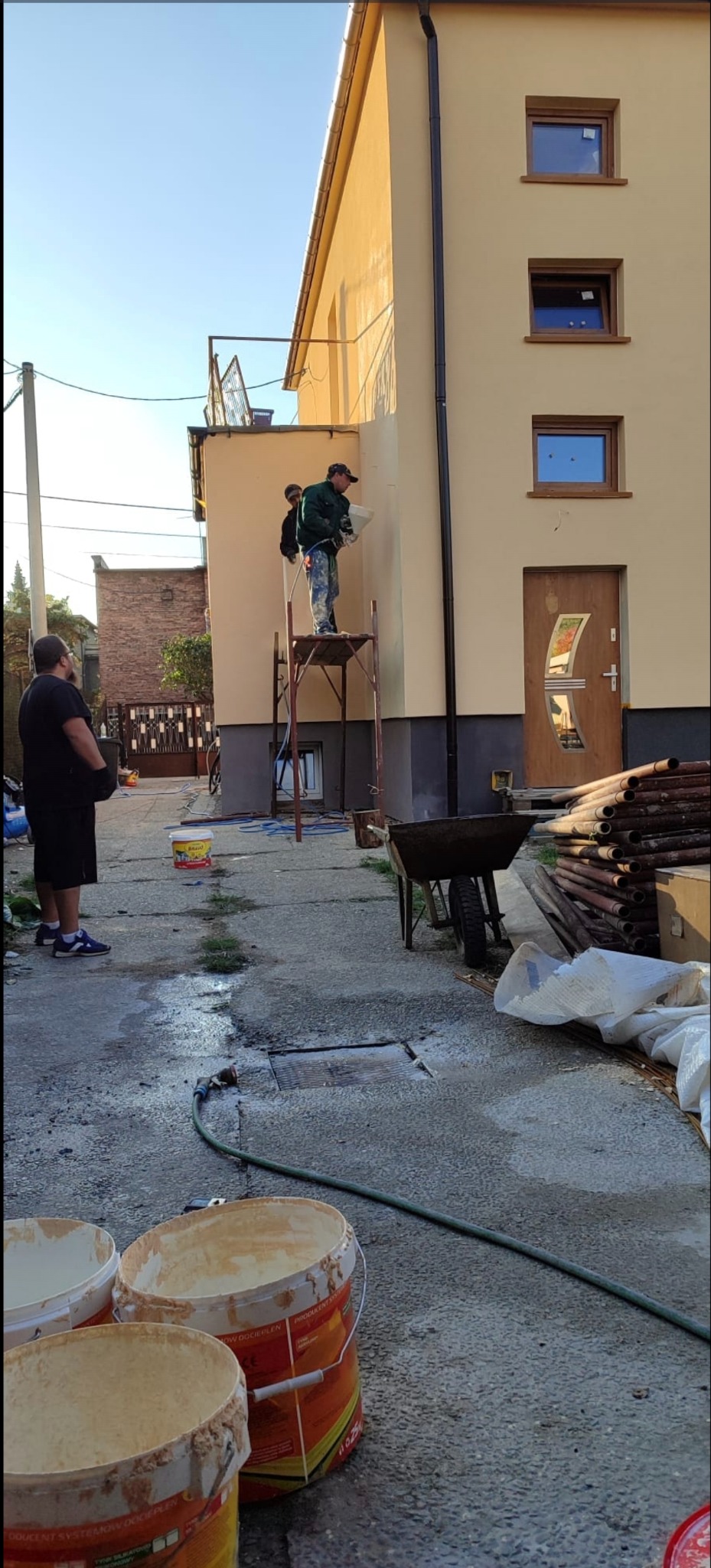Dwóch pracowników na rusztowaniu aplikuje tynk na elewację budynku. Widoczne wiaderka z zaprawą, rury i wózek. Jasna elewacja, brązowe okna i drzwi.