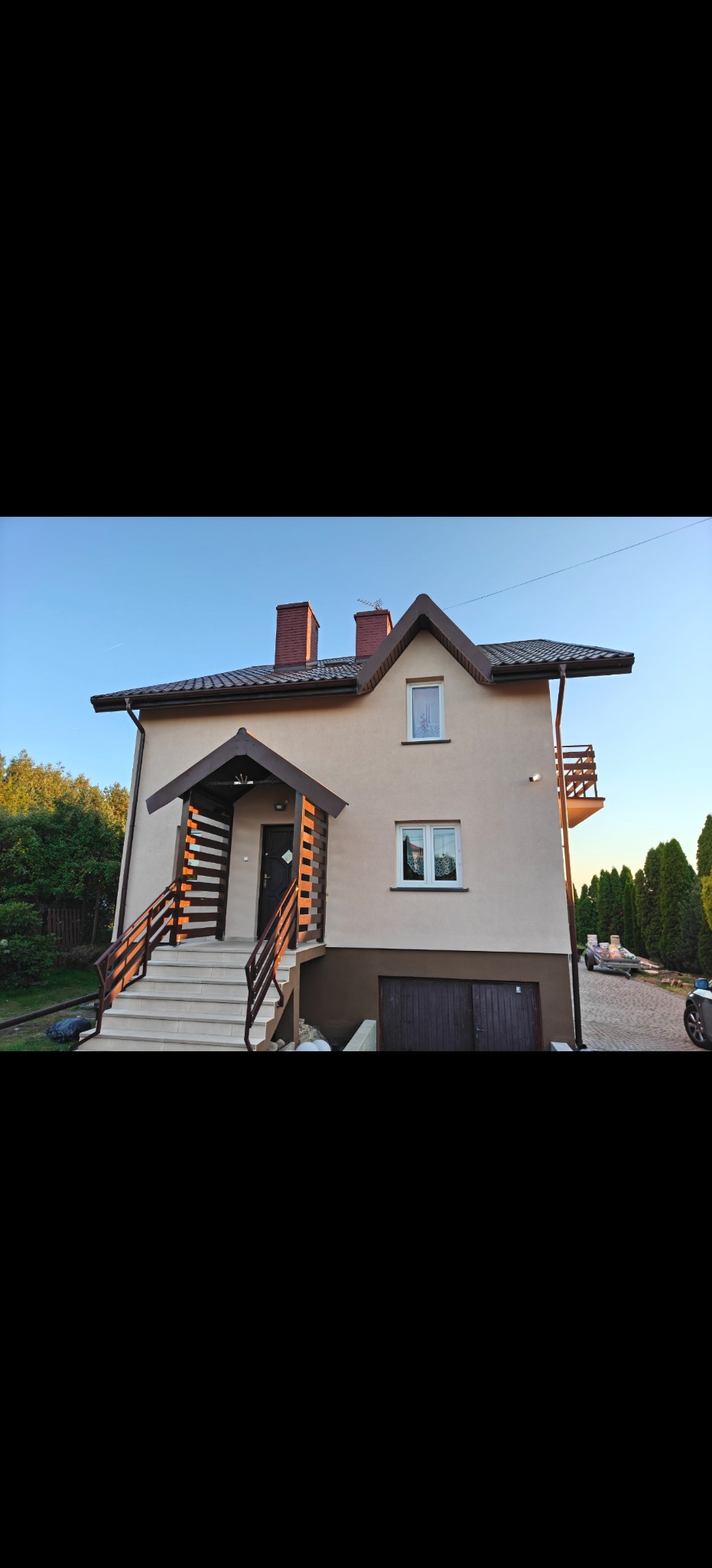 Dwukondygnacyjny dom z beżową elewacją, brązowym dachem i drewnianymi elementami wykończeniowymi, otoczony zielenią. Widoczny garaż i balkon.