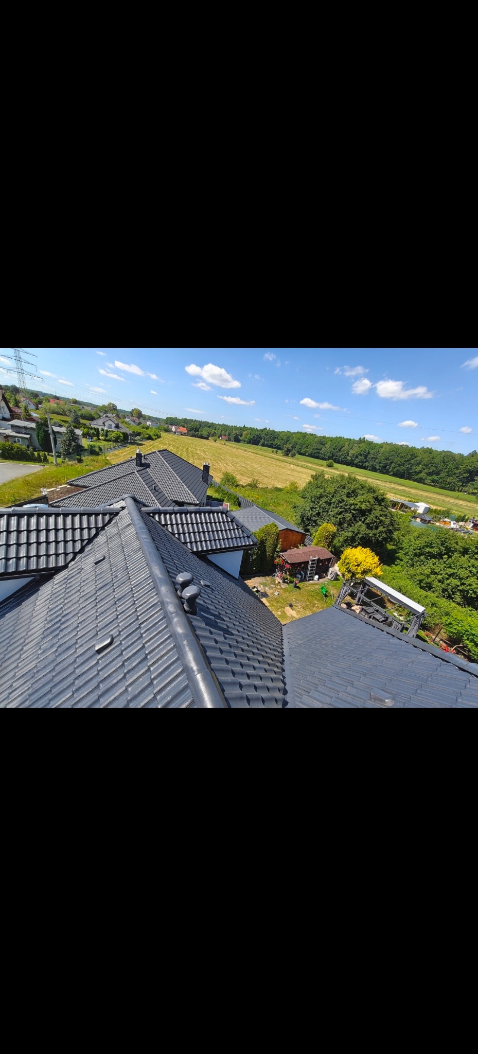Błyszczący, grafitowy dach z dachówki ceramicznej, widziany z góry na tle zieleni i błękitnego nieba. Widoczne kominy wentylacyjne i elementy wykończenia dachu.