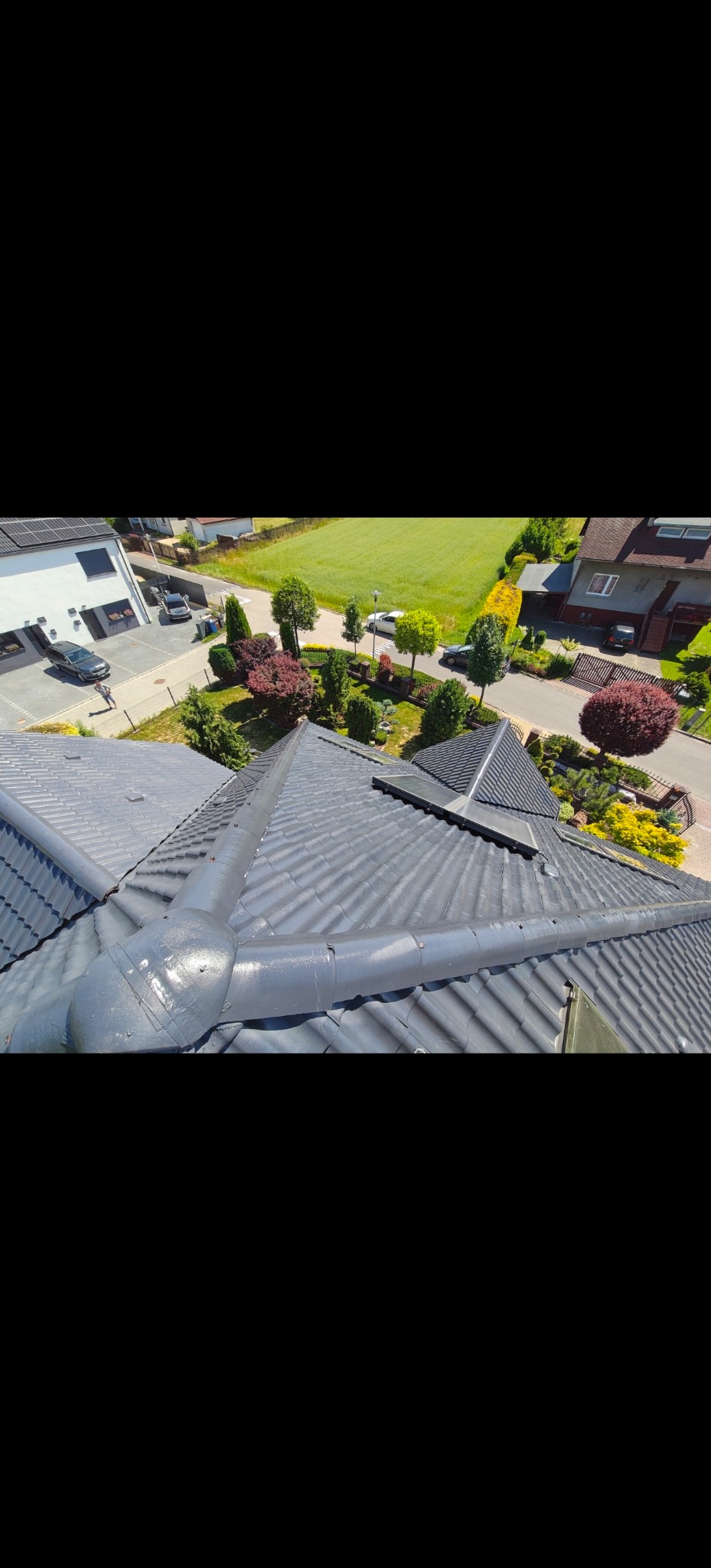 Szary, pomalowany dach z dachówki ceramicznej, widok z góry na tle zieleni i zabudowy jednorodzinnej w słoneczny dzień. Widoczne detale łączeń i faktura dachówki.