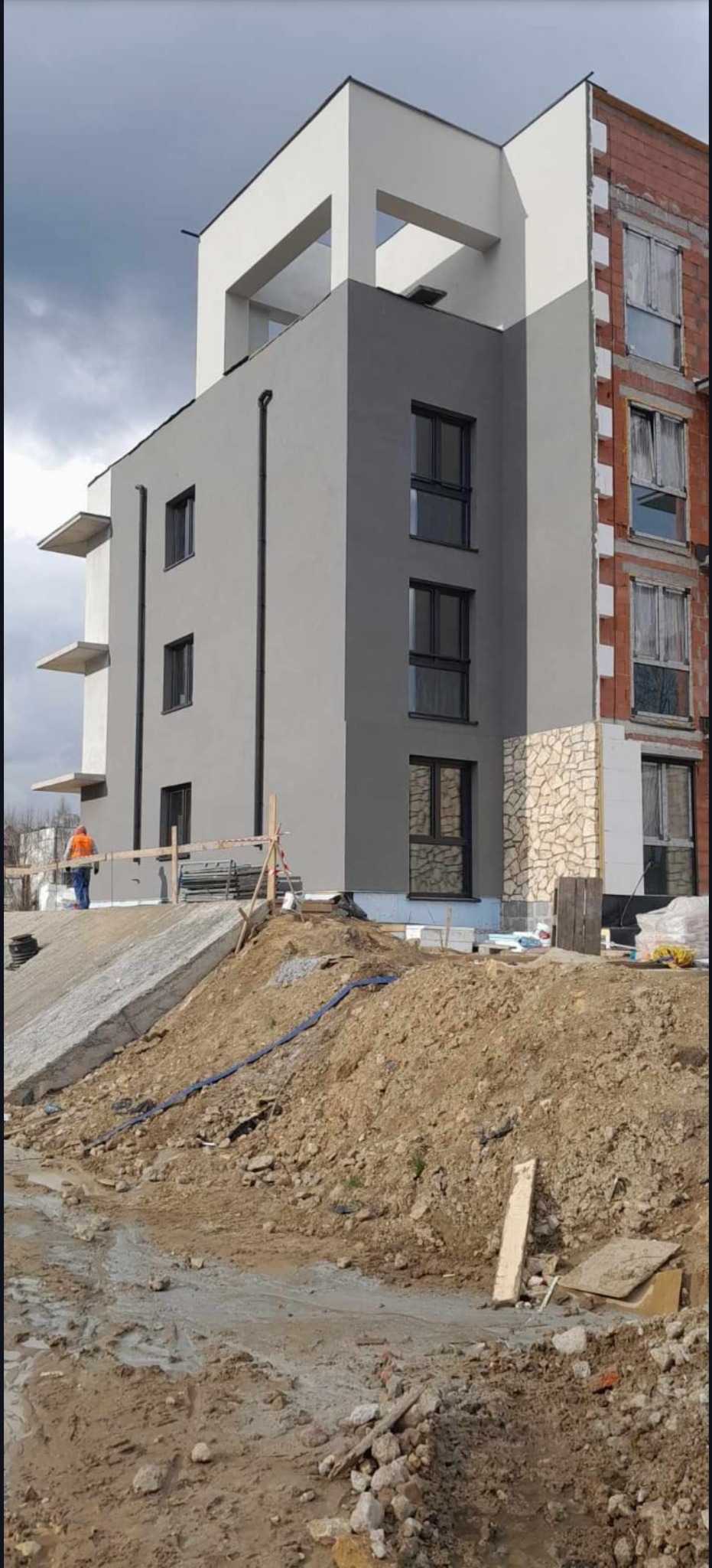 Wielopiętrowy budynek po częściowym malowaniu elewacji na szaro, z fragmentami w stanie surowym i osobą w oddali. Widoczne prace ziemne.