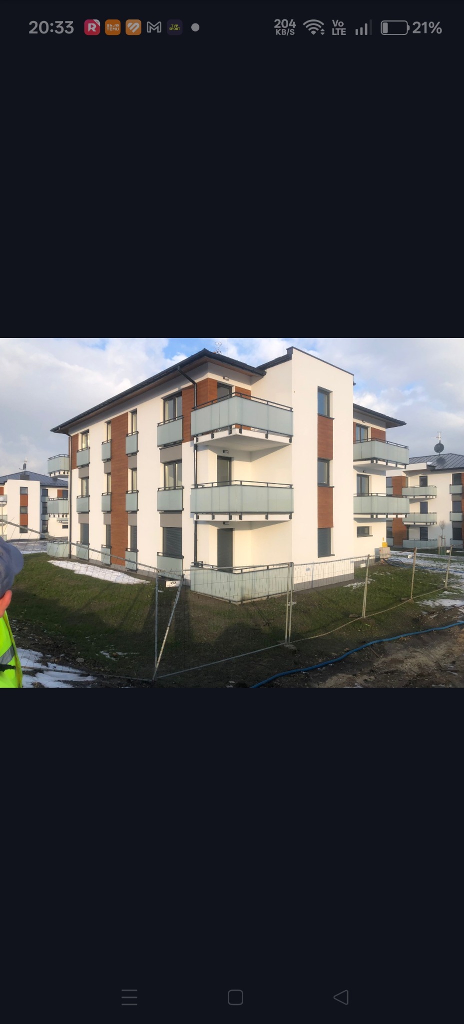 Nowoczesny, trzypiętrowy budynek mieszkalny z balkonami i elementami drewnopodobnymi na elewacji, otoczony trawnikiem i tymczasowym ogrodzeniem.