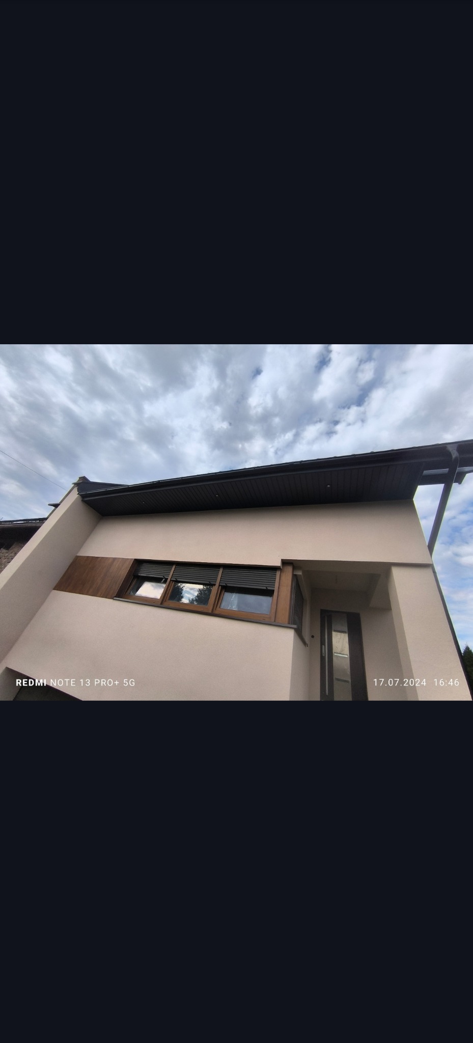 Elewacja domu jednorodzinnego po renowacji, z widocznymi oknami z drewnianą stolarką i świeżo pomalowaną fasadą w odcieniach beżu, sfotografowana w pochmurny dzień.