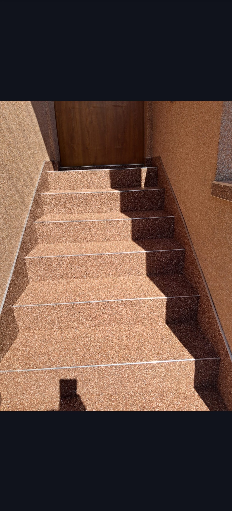 Schody wejściowe wykończone żywicą kamienną w odcieniach brązu, z metalowymi listwami krawędziowymi. Widoczny cień osoby fotografującej.