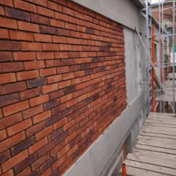Częściowo wykończona elewacja z cegły klinkierowej w odcieniach czerwieni i brązu, widoczna na tle szarego betonu i rusztowania.