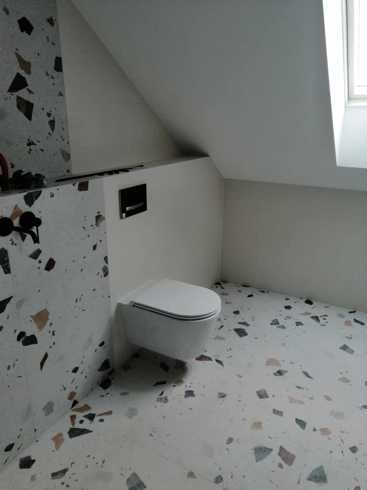 Nowoczesna łazienka na poddaszu z toaletą podwieszaną, wykończona płytkami imitującymi lastryko w odcieniach szarości, brązu i czerni, z czarną armaturą i minimalistycznym panelem sterującym spłuczką.