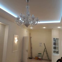 SLAVLEN GRUP Viacheslav Kudinov - Wykończone wnętrze pokoju z białym sufitem podwieszanym z oświetleniem LED, ozdobnym żyrandolem i ścianami w odcieniach beżu, widoczne drabina malarska i akcesoria malarskie.