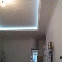 SLAVLEN GRUP Viacheslav Kudinov - Podwieszany sufit z oświetleniem LED w kształcie prostokąta, listwy dekoracyjne na ścianie i suficie, widoczna drabina i kartony w tle, surowy stan wykończenia wnętrza.